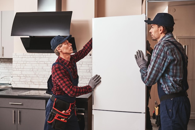 Снова переезжаем. мужчины механики переносят холодильник в другое место. Они помогают друг другу. Вид сбоку