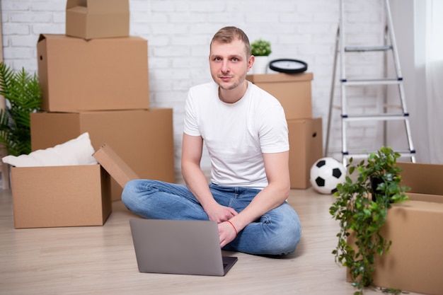 Moving day - young handsome man using laptop in new apartments surrounded with cardboard boxes