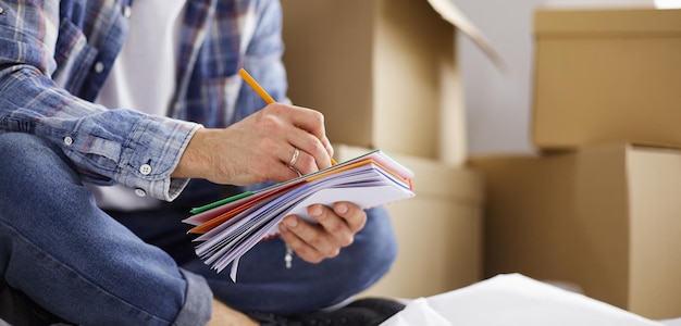 A moving man sitting on the floor in empty apartment Among the Boxes Checking the List of Things