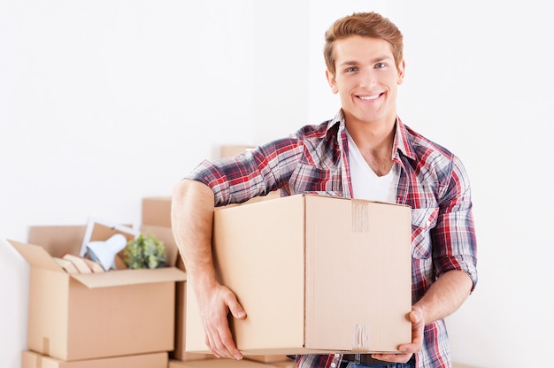 Moving to a new apartment. Cheerful young man holding a cardboard box and smiling while other carton boxes laying on background