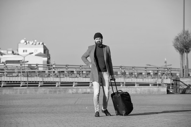 Moving to new city alone Hipster ready enjoy travel Carry travel bag Man bearded hipster travel with luggage bag on wheels Traveler with suitcase arrive airport railway station urban background