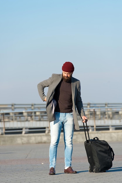 Один переезд в новый город. Путешественник с чемоданом прибывает на фоне городского железнодорожного вокзала аэропорта. Хипстер готов наслаждаться путешествиями. Носите дорожную сумку. Человек бородатый хипстер путешествует с багажной сумкой на колесах.
