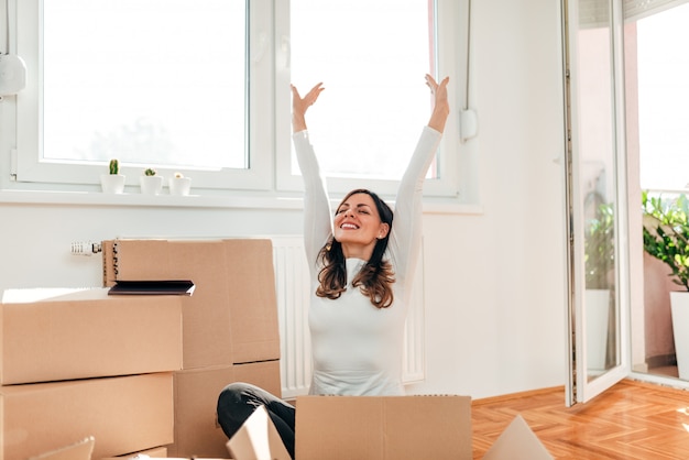 Moving to new home. Happy woman with arms raised sitting on the floor among carton boxes.