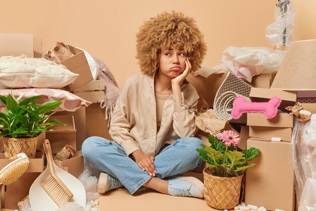 Moving to own home upset displeased woman sits in lotus pose on\
floor in empty room with cardboard boxes and plants in pots around\
bought new property feels lonely as lives alone relocation\
concept