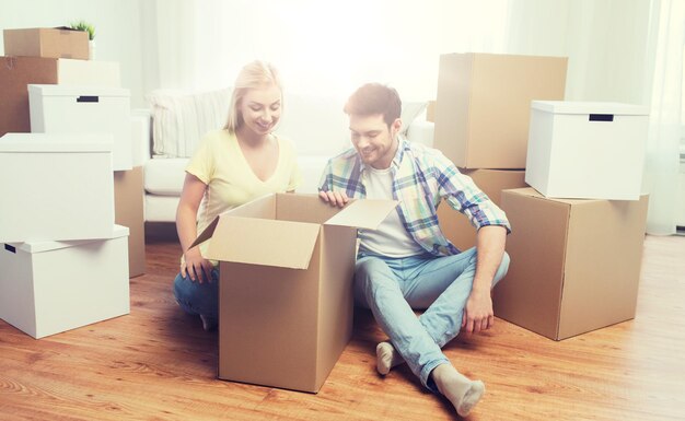 Photo moving, people, repair and real estate concept - smiling couple looking into big cardboard box at new home