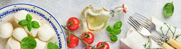 Mozzarella cheese with basil in white ceramic plate, tomato cherry olive oil and spice grinder, condiments, pepper tomato cherry on slate stone background. Mock up. Ingredients for Caprese salad.