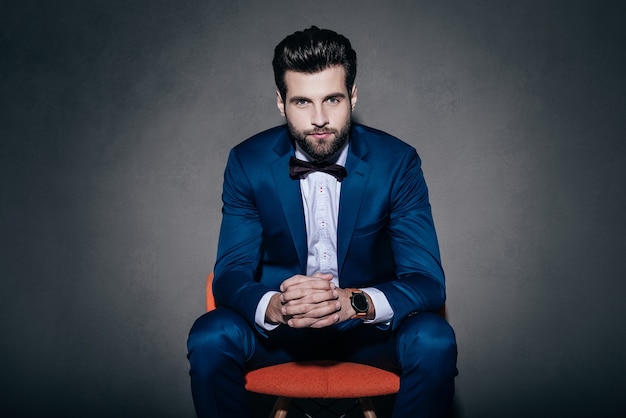 Mr. Confidence. Young handsome man in suit and bow tie looking at camera while sitting in orange chair against grey background