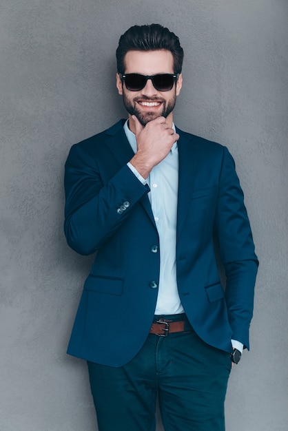 Mr. Handsome. Cheerful young handsome man in sunglasses keeping hand on chin and looking at camera with smile while standing against grey background