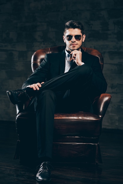 Photo mr. perfection.young handsome man in suit and sunglasses holding hand on chin and looking at camera while sitting in leather chair against dark grey background