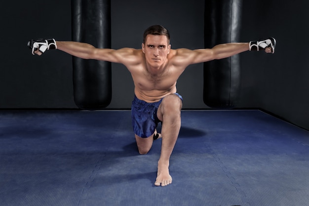 Muay thai-vechter in zwarte handschoenen en blauwe korte broek poseren tegen twee bokszakken in de sportschool.