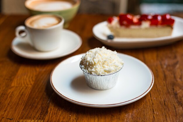 Muffin met stuk zelfgemaakte aardbeientaart en twee kopjes cappuccino met latte art op houten achtergrond