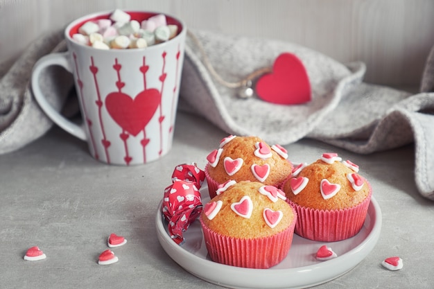 Muffin decorati con cuori di zucchero e una tazza con cuore rosso sul muro grigio chiaro