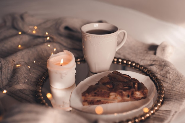 Mug of black tea with burning candle and chocolate pie in bed over Christmas lights close up. Good morning. Winter holiday season. Snack time.