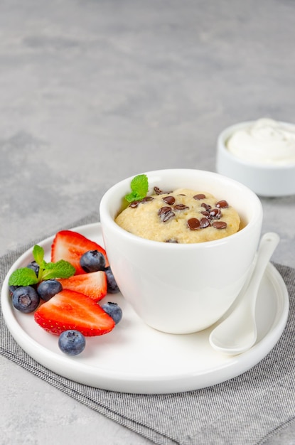 Mug cake with chocolate chips fresh berries and cream on a gray concrete background Copy space