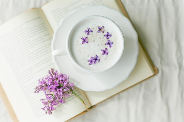Mug of cappuccino on a beige surface. Lilac flowers, book. 