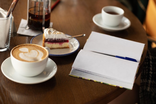 A mug of cappuccino a slice of cake and a notebook on the table in a cafe