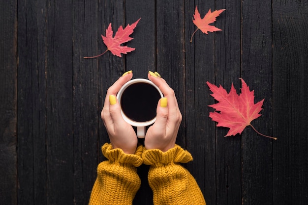 Mug of coffee in a female hand Wooden background autumn leaves Top view