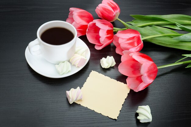 Mug of coffee, marshmallows, blank postcard and pink tulips. Black background.