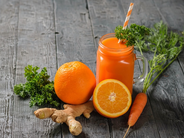 A mug of fresh carrot smoothie with cocktail straw, parsley, carrots, ginger root and oranges on a black table.