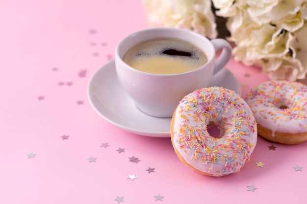 Mug of hot cappuccino and two donuts with flowers on pink