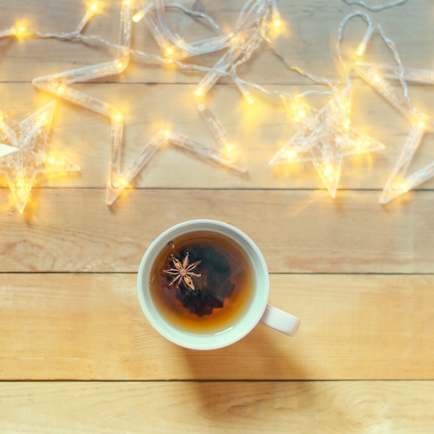 A mug of hot tea with spices on a wooden background with Christmas garland