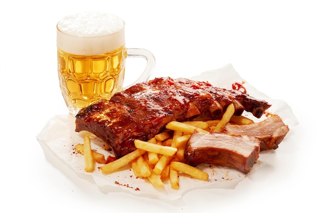 Mug of refreshing beer and plate with barbeque pork ribs and French fries placed on white table in studio