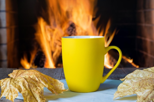 Mug of tea or coffee and autumn leaves before cozy fireplace in country house