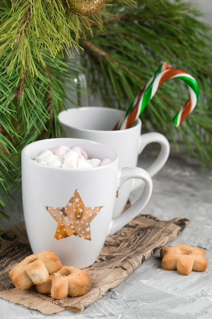 Mug with drink decorated with marshmallow and star shape cookies  near evergreen christmas tree branches gray concrete background