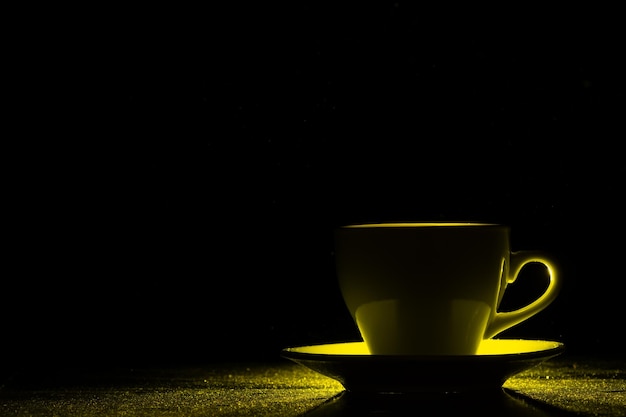 A mug with a warm drink illuminated by yellow light on black background Morning coffee