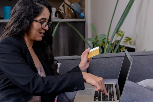 Mujer 라티 haciendo compras en su computadora portatil y pagando con tarjeta de credito desde casa