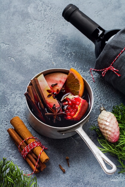 Mulled wine hot drink with citrus, apple, pomegranate and spices in aluminum casserole with vintage hristmas tree toys and Fir branch on concrete surface. Selective focuse.