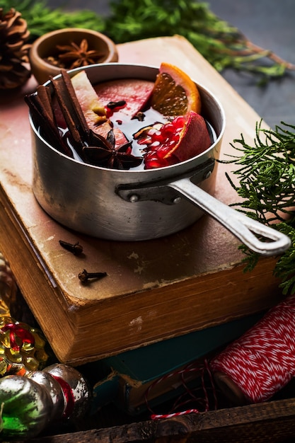 Mulled wine hot drink with citrus, apple, pomegranate and spices in aluminum casserole with vintage hristmas tree toys and Fir branch on concrete surface. Selective focuse.