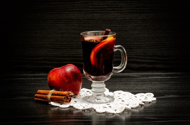 Mulled wine with spices on a lace napkin, apple and cinnamon sticks. Black wood background