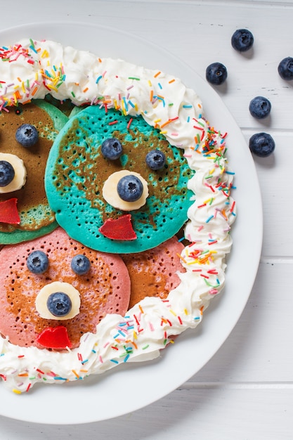 白い皿の上の子供のための果物とホイップクリームとマルチカラーのパンケーキ。離乳食用の目と口でパンケーキ。