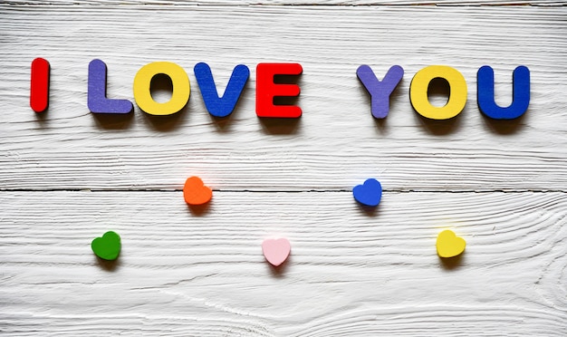 Multi-colored wooden letters making up the inscription I love you and multi-colored wooden hearts on a white wooden background. St. Valentine's Day background.