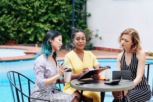 Multi-ethic business team of young serious women sitting at table by swimming pool, drinking coffee and discussing reports