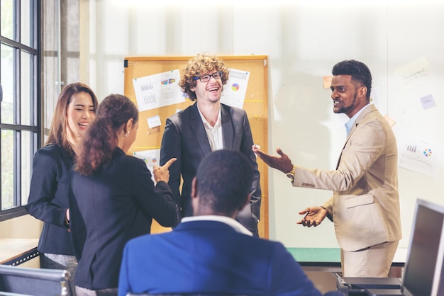 Squadra multietnica di affari vari impiegati che conversano e sorridono mentre tengono un incontro con i colleghi in un ufficio moderno, uomo d'affari anziano che fa una presentazione sullo sfondo