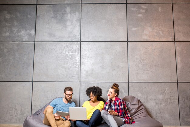 Multi ethnic coworkers dressed casually in colorful clothes working with different gadgets sitting on the bag chairs on the gray wall background. Wide shot with copy space