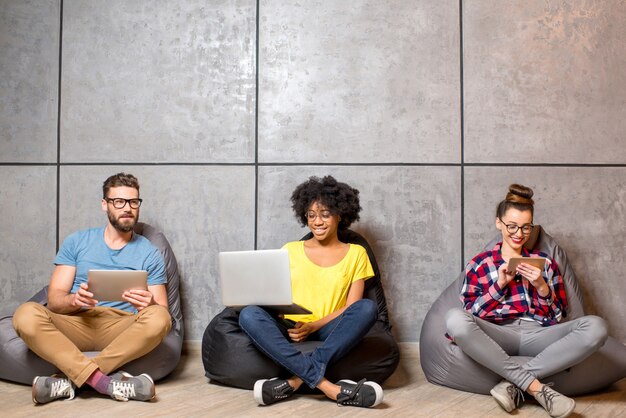 Multi ethnic coworkers dressed casually in colorful clothes working with different gadgets sitting on the bag chairs on the gray wall background