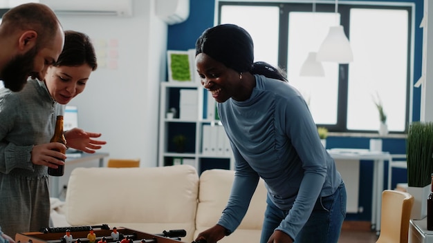 Multi ethnic coworkers playing soccer game at foosball table with drinks after work. Cheerful workmates enjoying football play, beer and snacks to celebrate party at office after hours