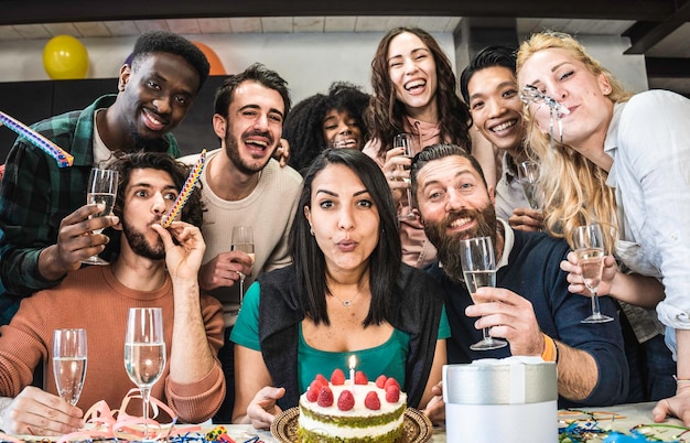 Foto amici multietnici che si divertono durante la festa di compleanno giovani che guardano la macchina fotografica durante la festa di compleanno per foto di gruppo ragazza marocchina che spegne la candela sulla torta prima di scartare il suo regalo