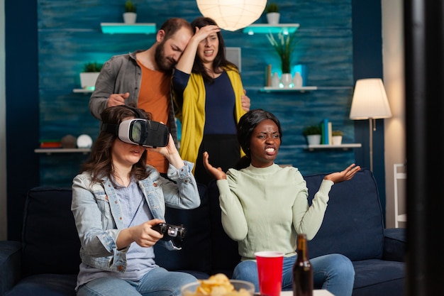 Multi ethnic friends losing online video games compettion experiencing virtual reality with headset. Mixed race group of people hanging out together having fun late at night in living room.