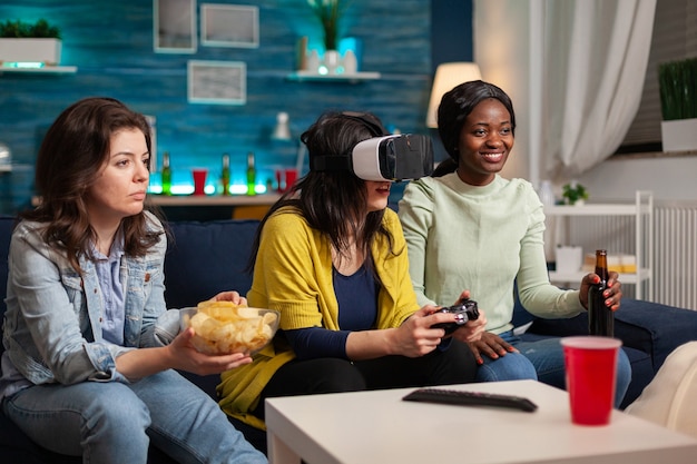 Multi ethnic friends socialising playing video games with vr goggles, drinking beer sitting on couch. Mixed race group of people hanging out together having fun late at night in living room.