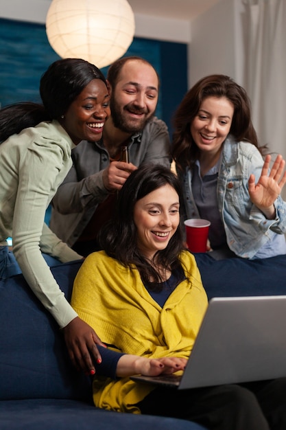 Photo multi ethnic friends waving at laptop webcam during video call sitting on couch late at night. group of multiracial people spending time together sitting on couch late at night in living room.