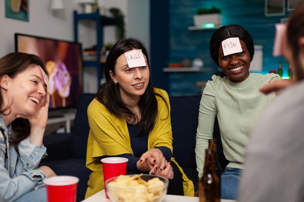 Multi-ethnic friends with stickey notes on forehead playing guess who game enjoying spending time together hanging out at home in living room. Group of people having fun during evening party