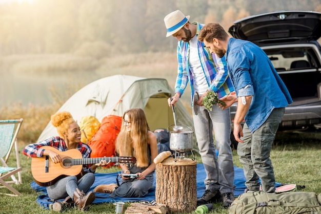 Multi etnico gruppo di amici vestiti casualmente facendo un picnic, cucinando zuppa con calderone durante la ricreazione all'aperto con tenda, auto e attrezzatura da trekking vicino al lago