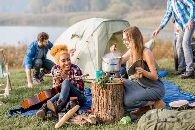 Multi ethnic group of friends dressed casually having a picnic, cooking soup with cauldron during the outdoor recreation near the lake