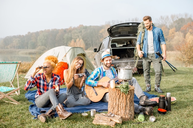 Multi ethnic group of friends dressed casually having a picnic during the outdoor recreation with tent, car and hiking equipment near the lake