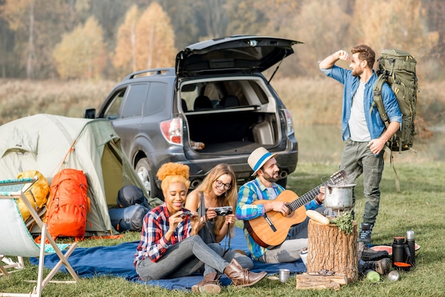 Многонациональная группа друзей, одетых в повседневную одежду, устраивает пикник во время отдыха на природе с палаткой, автомобилем и походным снаряжением возле озера