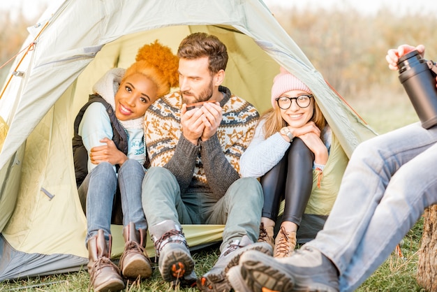 Multi ethnic group of friends dressed in sweaters warming up together sitting in the tent during the outdoor recreation near the lake